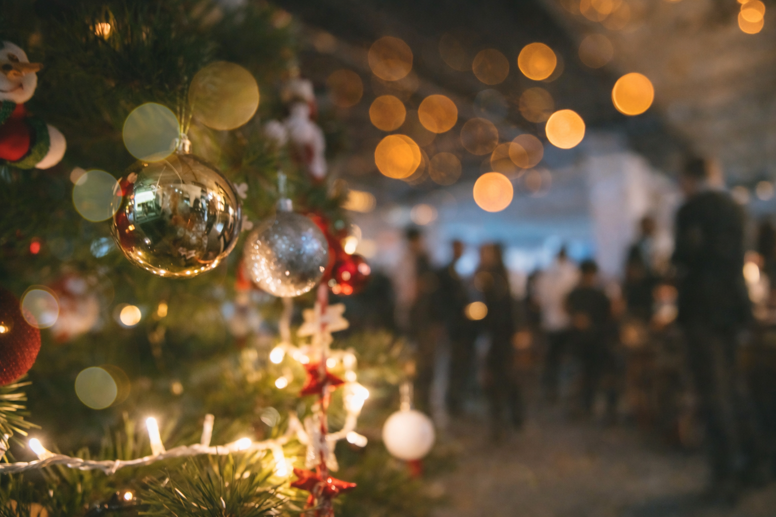 Vánočně ozdobený strom v popředí a rozostřené postavy lidí v pracovním prostředí v pozadí